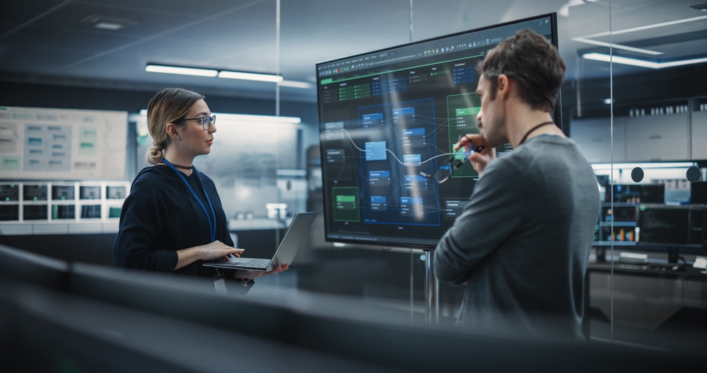 Two individuals standing and looking a TV/screen, engrossed in the content displayed on the screen.